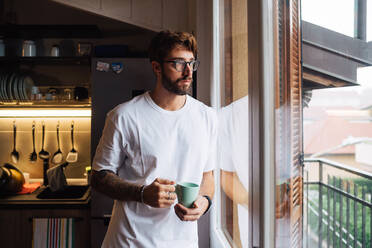 Mittlerer erwachsener Mann mit Kaffeetasse, der durch ein Wohnungsfenster hinausschaut - ISF21457