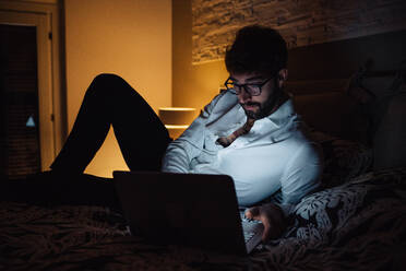 Mittlerer erwachsener Mann, der nachts auf dem Bett liegt und einen Laptop benutzt - ISF21467