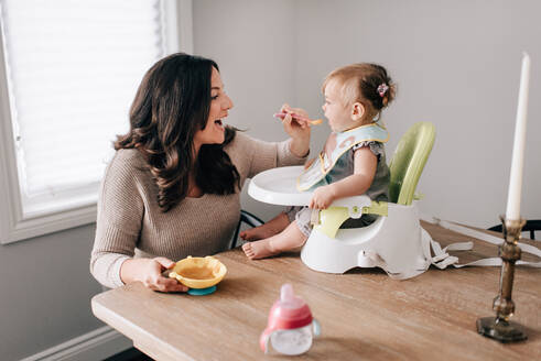 Mother feeding baby daughter in child seat on kitchen table - ISF21475