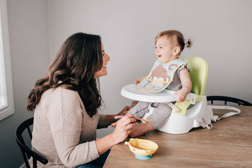 Mother feeding baby daughter in child seat on kitchen table - ISF21476