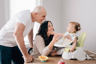 Mother and father feeding baby daughter in child seat on kitchen table - ISF21477