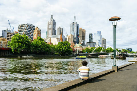 Mann sitzt am Yarra-Fluss und schaut auf Wolkenkratzer in Melbourne, Victoria, Australien, lizenzfreies Stockfoto