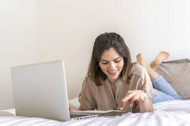 Happy young woman lying on bed using smartphone and laptop - AFVF03292