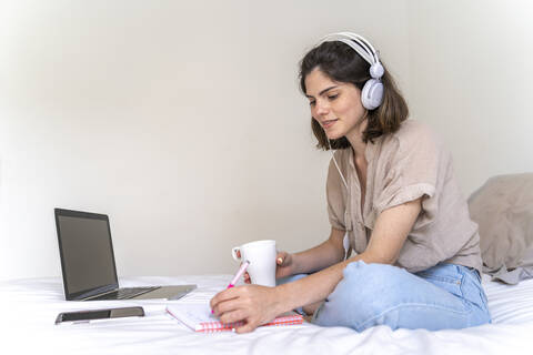 Junge Frau sitzt mit Kopfhörern und Laptop auf dem Bett und macht sich Notizen, lizenzfreies Stockfoto