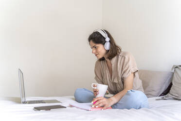 Junge Frau sitzt mit Kopfhörern und Laptop auf dem Bett und macht sich Notizen - AFVF03294