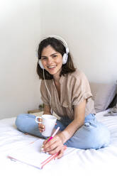 Porträt einer glücklichen jungen Frau, die mit Kopfhörern auf dem Bett sitzt und Notizen macht - AFVF03295