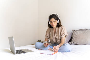 Junge Frau sitzt mit Kopfhörern und Laptop auf dem Bett und macht sich Notizen - AFVF03296