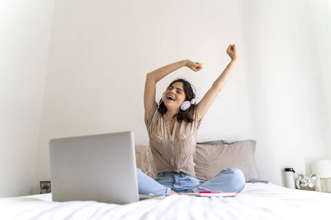 Glückliche junge Frau sitzt mit Kopfhörern und Laptop auf dem Bett, lizenzfreies Stockfoto