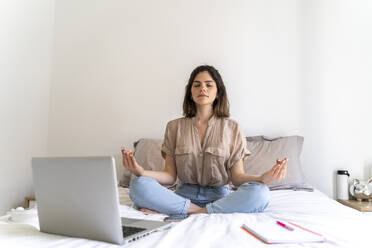 Young woman sitting on bed with laptop practicing yoga - AFVF03299