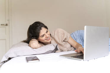 Glückliche junge Frau auf dem Bett liegend mit Laptop - AFVF03306