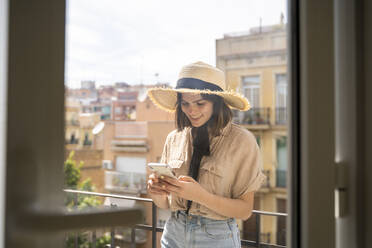 Smiling young woman on balcony in the city wearing straw hat using cell phone - AFVF03314