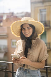 Portrait of smiling young woman wearing straw hat standing on balcony in the city - AFVF03315