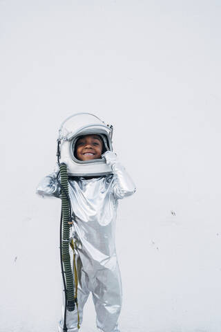 Smiling little girl wearing space suit putting on space hat in front of white background stock photo