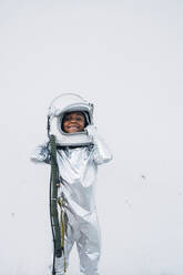 Smiling little girl wearing space suit putting on space hat in front of white background - JCMF00069