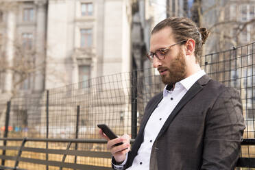 Bärtiger junger Geschäftsmann mit Brille, der auf sein Handy schaut, New York City, USA - MFRF01302