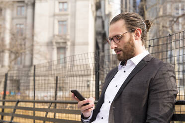 Bearded young businessman wearing glasses looking at cell phone, New York City, USA - MFRF01302