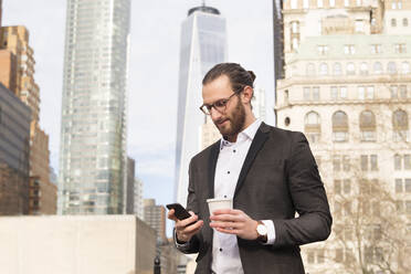 Bärtiger junger Geschäftsmann mit Kaffee zum Mitnehmen schaut auf sein Handy, New York City, USA - MFRF01303