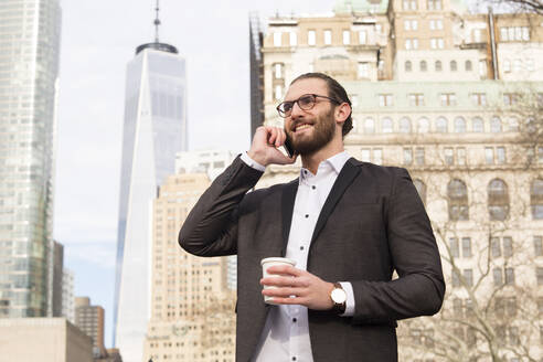 Lächelnder junger Geschäftsmann mit Kaffee zum Mitnehmen am Telefon, New York City, USA - MFRF01304