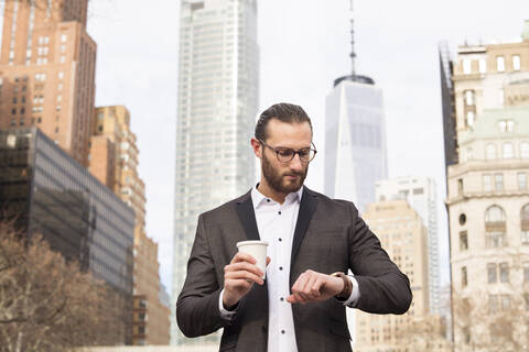 Bärtiger junger Geschäftsmann mit Kaffee zum Mitnehmen, der die Zeit überprüft, New York City, USA, lizenzfreies Stockfoto