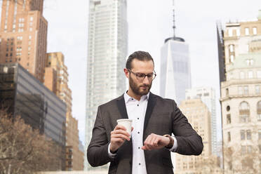 Bearded young businessman with coffee to go checking the time, New York City, USA - MFRF01305