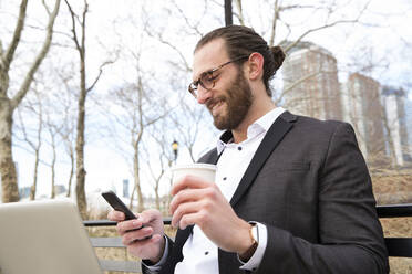 Smiling young businessman with coffee to go looking at cell phone, New York City, USA - MFRF01310
