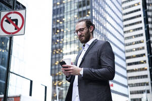 Bärtiger junger Geschäftsmann mit Kaffee zum Mitnehmen schaut auf sein Handy, New York City, USA - MFRF01311