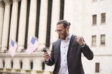 Aufgeregter junger Geschäftsmann, der auf sein Handy schaut, vor der Börse, New York City, USA - MFRF01318