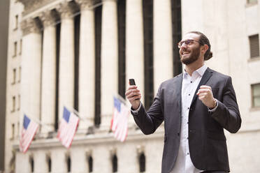 Glücklicher junger Geschäftsmann mit Mobiltelefon vor der Börse, New York City, USA - MFRF01320