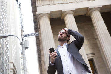Portrait of worried young businessman with cell phone in front of Stock Exchange, New York City, USA - MFRF01324