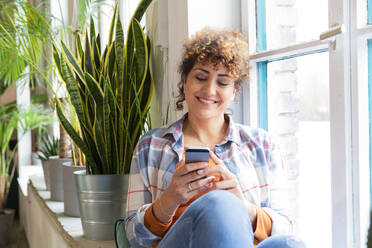 Smiling woman sitting at the window in office using cell phone - FKF03378