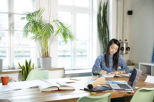 Frau arbeitet am Tisch in einem modernen Büro und macht sich Notizen - FKF03403