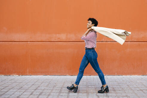 Businesswoman walking at a red wall in the background, putting her jacket on her shoulder - JRFF03283