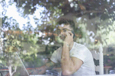 Young man with laptop on cell phone behind windowpane of a cafe - JPTF00160