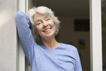 Portrait of smiling mature woman standing in front of open terrace door - PNEF01684