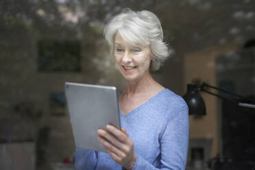 Porträt einer lächelnden reifen Frau hinter einer Fensterscheibe mit Blick auf ein digitales Tablet - PNEF01688