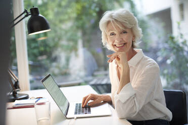 Portrait of smiling mature businesswoman sitting at desk using laptop - PNEF01730