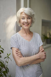 Portrait of smiling mature woman standing in front of opened terrace door - PNEF01732