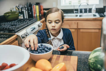 Gemischtrassiges Mädchen pflückt Blaubeeren aus einer Schüssel in der Küche - BLEF07788