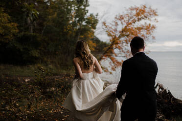 Romantic young groom carrying bride's wedding dress train on lakeside, rear view - ISF21515