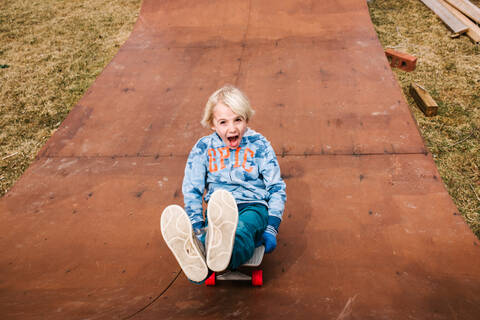 Junge sitzt und fährt rückwärts eine hölzerne Skateboard-Rampe hinunter, Porträt, lizenzfreies Stockfoto