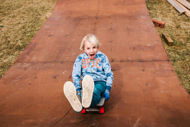 Boy sitting and skateboarding backwards down wooden skateboard ramp, portrait - ISF21546