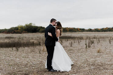 Romantic young bride and groom hugging in field, full length - ISF21561