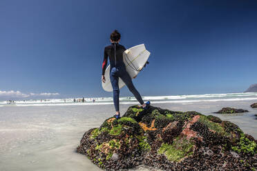 Junge Surfer, der auf einem Felsen am Strand steht und aufs Meer hinausschaut, Kapstadt, Westkap, Südafrika - ISF21585