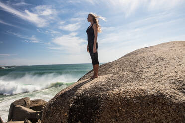 Junge Frau, die Yoga praktiziert, macht eine Pause und schaut von einem Felsen am Strand aus, Kapstadt, Westkap, Südafrika - ISF21593