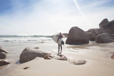 Young female surfer carrying surfboard looking out at ocean waves from beach, full length, Cape Town, Western Cape, South Africa - ISF21601