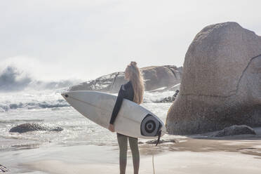 Young female surfer carrying surfboard looking out at ocean waves from beach, Cape Town, Western Cape, South Africa - ISF21602