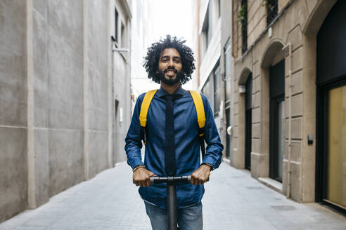 Porträt eines lächelnden jungen Mannes mit Rucksack auf einem E-Scooter nach der Arbeit, Barcelona, Spanien - JRFF03360