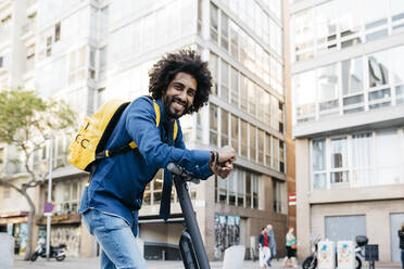 Porträt eines lächelnden jungen Mannes mit gelbem Rucksack auf einem E-Scooter in der Stadt, Barcelona, Spanien - JRFF03363