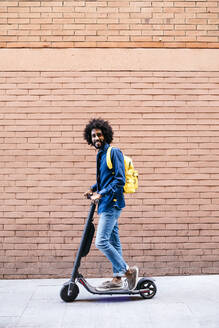 Portrait of smiling young man on E-Scooter in front of brick wall - JRFF03365