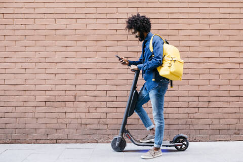 Junger Mann mit Rucksack und E-Scooter steht vor einer Backsteinmauer und schaut auf sein Handy, lizenzfreies Stockfoto